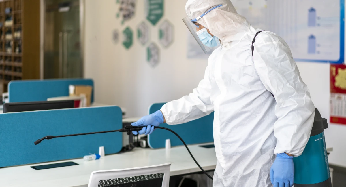 A cleaning staff member, wearing a full white protective suit, face shield, and gloves, is using a sprayer to perform deep disinfection and cleaning inside an office to prevent the spread of the virus.